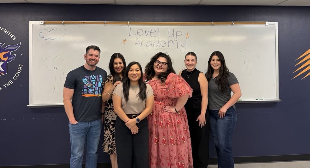 Six New Pathways for Youth staff members pose together in front of a whiteboard labeled 'Level Up Academy' during a team gathering.